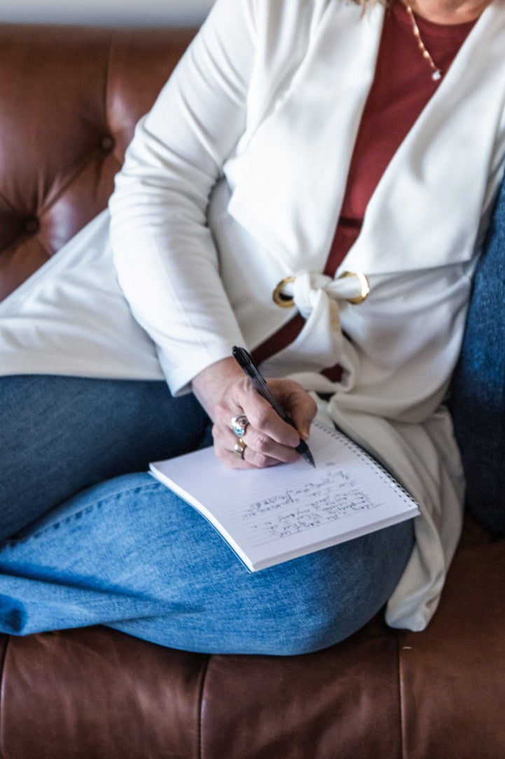 Woman sitting on a brown leather couch reading a piece of paper, with a colorful painting and plant in the background.