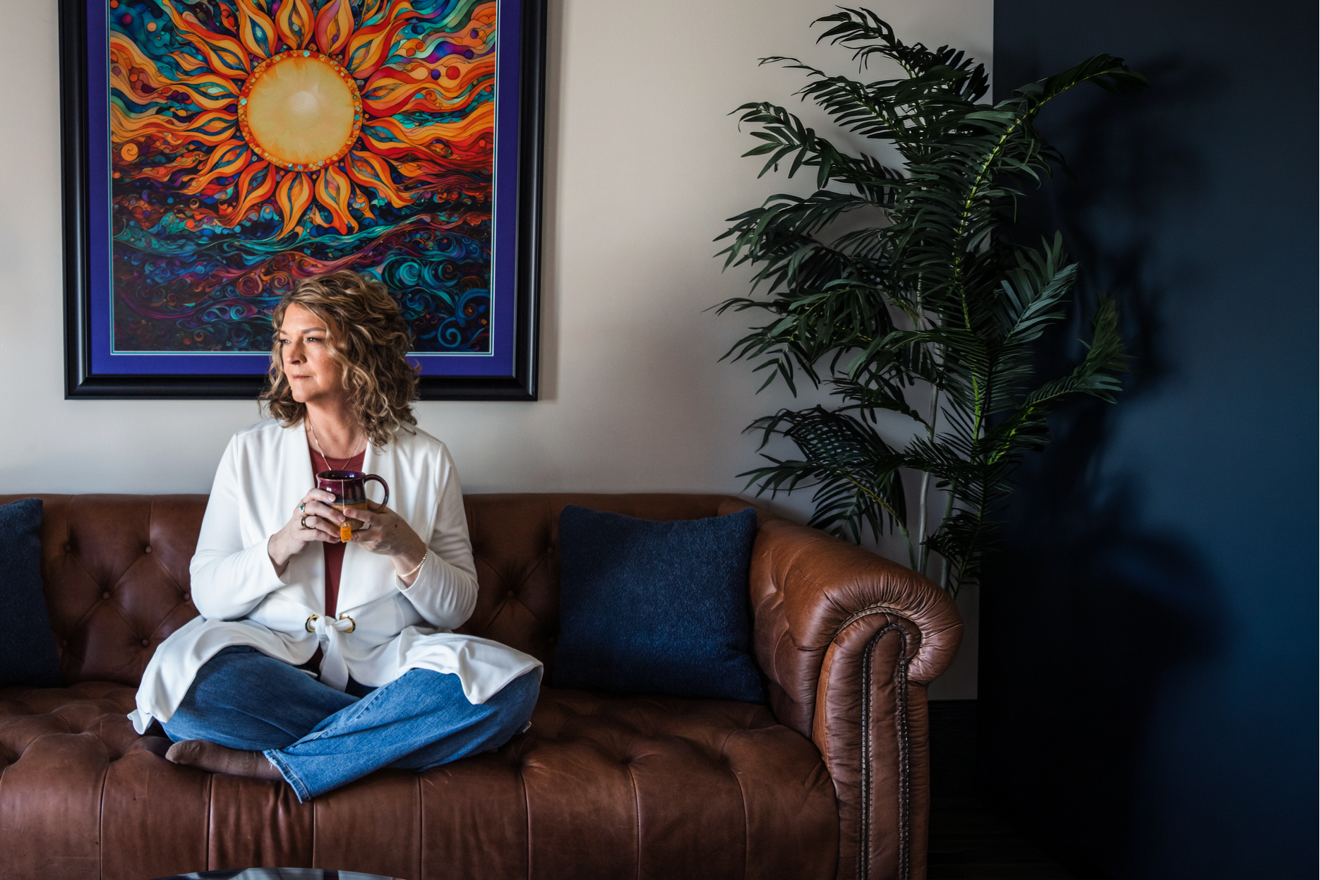 Woman sitting on a brown leather couch in a living room with a colorful painting of a sun on the wall.